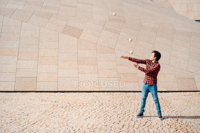 Joven hombre capacitado en camisa a cuadros realizando truco con bolas malabares mientras está de pie contra la estructura de hormigón contemporáneo en la calle urbana - foto de stock