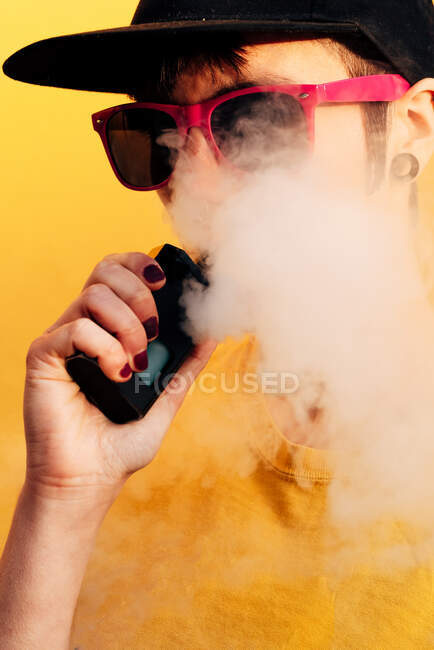 Contemporary female in stylish outfit exhaling fume while standing near yellow wall and vaping on city street — Stock Photo