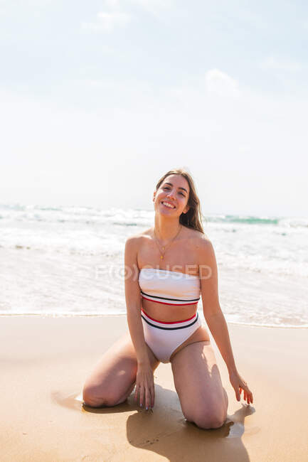 Улыбающаяся молодая женщина в купальнике сидит на песчаном пляже и смотрит в камеру возле пенного океана под голубым небом при дневном свете — стоковое фото