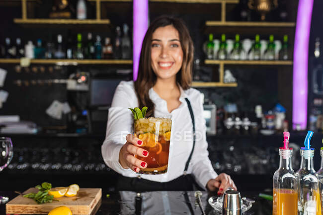 Щаслива молода жіночий бармен у стильному одязі, яка дивиться на камеру, подаючи коктейль мохіто зі шматочками лимона, стоячи за прилавком у сучасному барі. — стокове фото