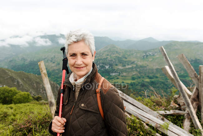 Smiling elderly woman with backpack and trekking stick standing on grassy slope towards mountain peak during trip in nature looking at camera — Stock Photo