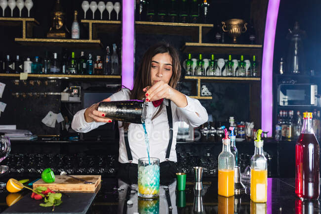 Barkeeperin in stylischem Outfit serviert blauen Cocktail aus einem Shaker in ein Glas, während sie an der Theke in einer modernen Bar steht — Stockfoto