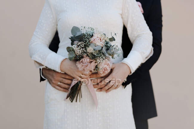 Crop marié anonyme embrassant mariée élégante en robe de mariée blanche avec bouquet floral délicat — Photo de stock
