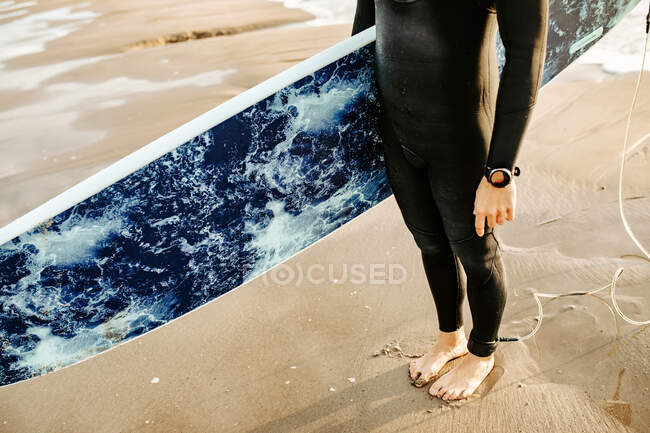 Side view of cropped unrecognizable surfer man dressed in wetsuit standing with the surfboard on the water waiting to catch a wave on the beach during sunrise — Stock Photo