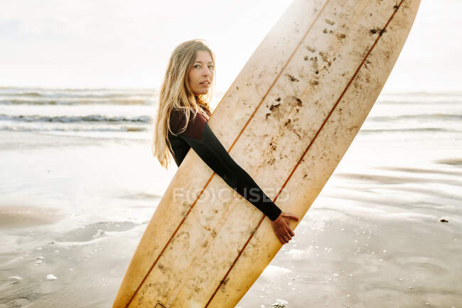 Vista lateral de la mujer surfista vestida con traje de neopreno de pie mirando a la cámara con la tabla de surf en la playa durante el amanecer en el fondo - foto de stock