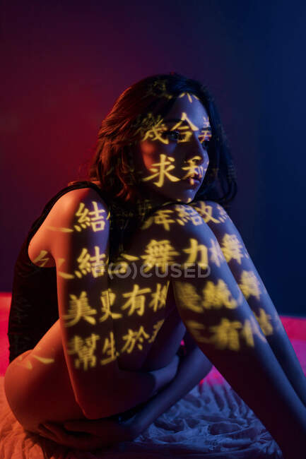 Fashionable young female model with light projection in shape of oriental hieroglyphs looking away sitting leaning on knees in dark studio with red illumination — Stock Photo