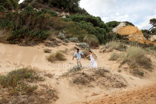 Deleitado puente multirracial y novio tomados de la mano y corriendo a lo largo de la colina de arena en el día de la boda en la naturaleza - foto de stock