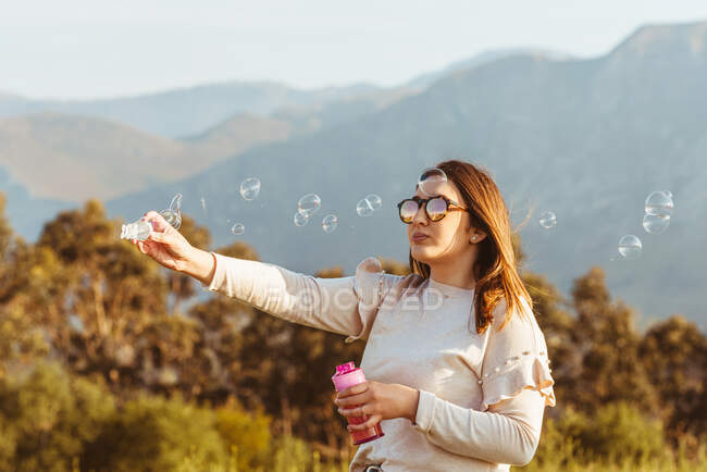 Content stylish female in sunglasses holding stick and making soap bubble in wind standing on meadow in mountains — Stock Photo