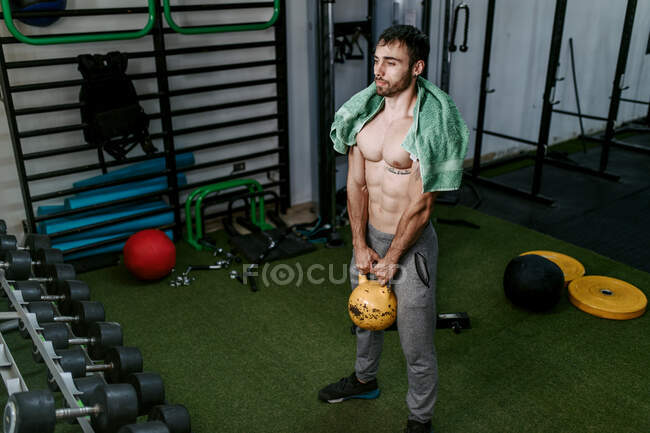 Atlético masculino com tronco nu fazendo exercícios com kettlebell pesado durante o treinamento ativo no centro esportivo — Fotografia de Stock