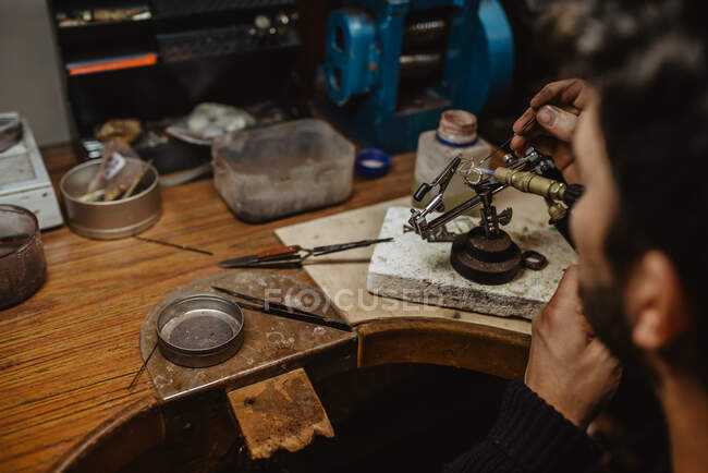 Orfebre anónimo que usa soplete para calentar diminutos adornos metálicos mientras hace joyas en el banco de trabajo - foto de stock