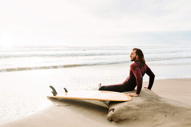 Vista lateral do surfista pensativo vestido de fato de mergulho sentado com prancha de surf na praia durante o nascer do sol — Fotografia de Stock
