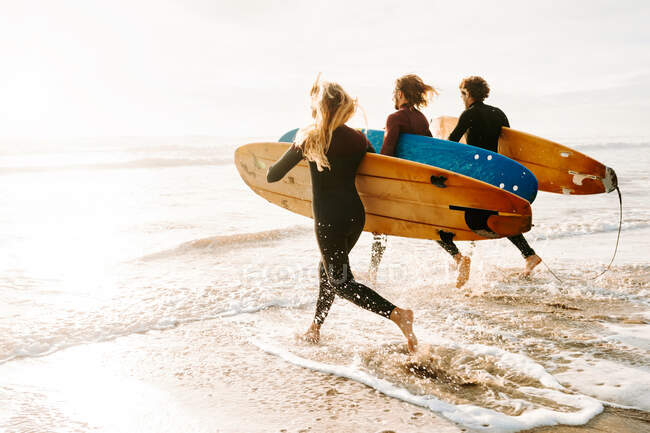Side view of group of surfer friends dressed in wetsuits running with surfboards towards the water to catch a wave on the beach during sunrise — Stock Photo