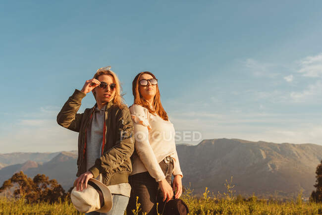 Giovani amiche vicine in abiti eleganti in piedi insieme sul prato in montagna guardando lontano in luce dorata — Foto stock