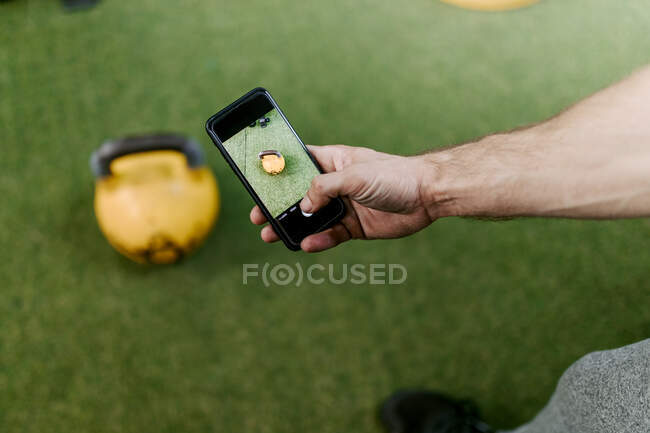 Zugeschnittene unkenntliche Person, die mit dem Smartphone ein Foto von der bunten Kettlebell macht, die auf dem Boden eines modernen Fitnesscenters platziert wurde — Stockfoto