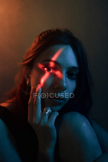 Jovem modelo feminino sem emoção com projeção de luz em forma de cruz no rosto sentado no estúdio escuro e olhando para a câmera — Fotografia de Stock