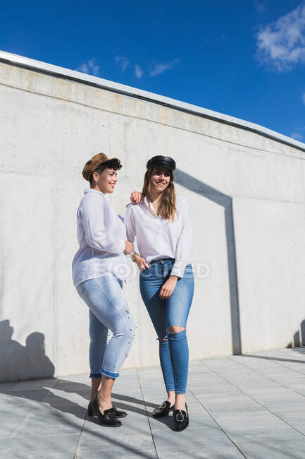 Full body of young positive female friends in trendy outfits and hats standing on walkway near gray wall in sunny day under blue sky — Stock Photo