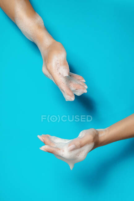 Cosecha femenina anónima con uñas largas demostrando manos con espuma de jabón blanco sobre fondo azul - foto de stock