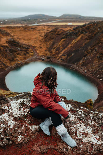 Desde arriba vista lateral de un joven turista sentado en el pico de la montaña y mirando el agua en el valle - foto de stock