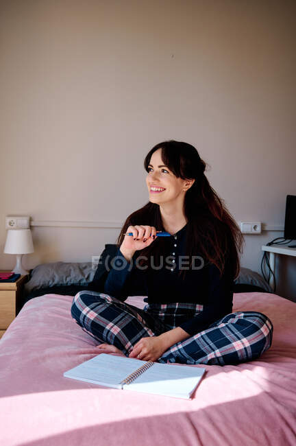 Молодая восторженная студентка сидит на кровати с блокнотом и делает домашнюю работу дома — стоковое фото