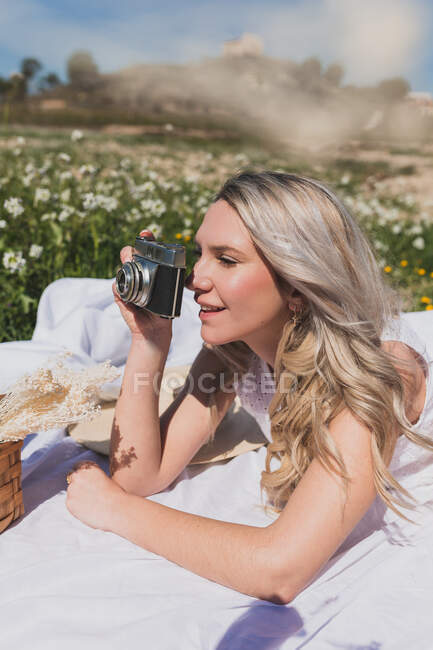 Mulher positiva deitada na xadrez e tirando foto na câmera à moda antiga no dia de verão no campo — Fotografia de Stock