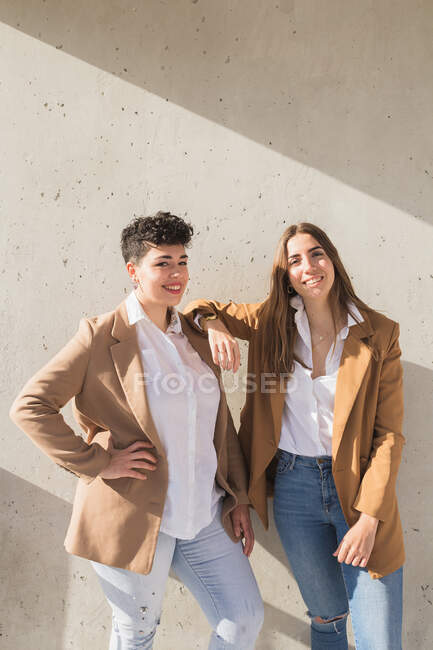 Jóvenes mujeres sonrientes con ropa elegante de pie juntos mientras miran a la cámara en un día soleado cerca de la pared gris - foto de stock