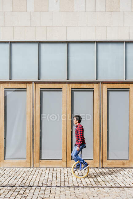 Vista lateral de cuerpo completo de hombre joven activo en camisa a cuadros y jeans realizar truco en monociclo cerca de la pared de vidrio espejado de edificio contemporáneo - foto de stock