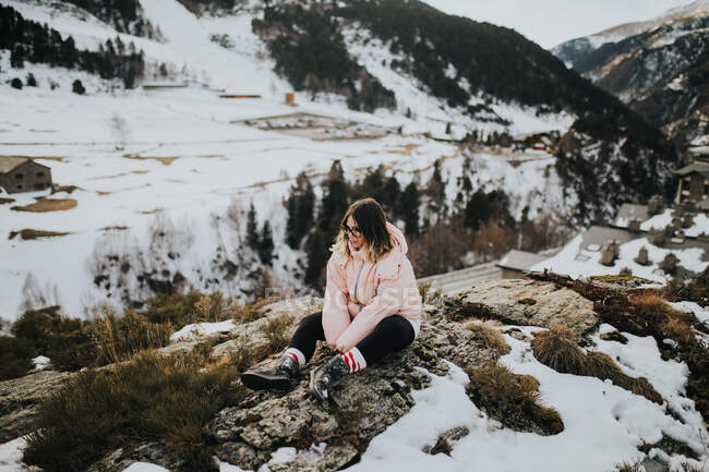 Young happy tourist in eyeglasses and sweater looking away and sitting between woods in snow near mountain — Stock Photo