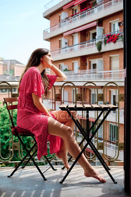 Vista laterale della graziosa femmina in abito estivo seduta a tavola sul balcone e guardando altrove — Foto stock