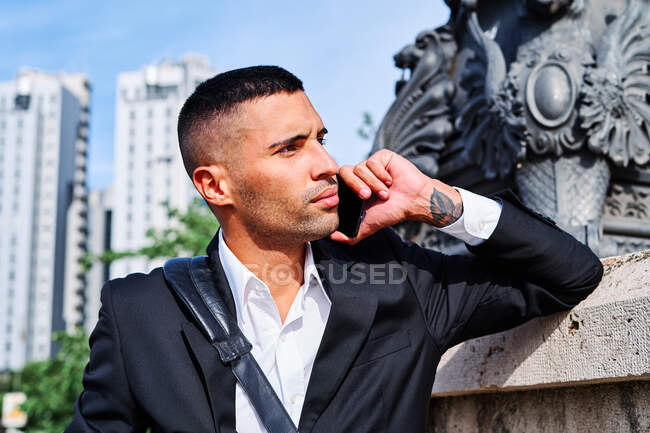 Full body Hispanic man in stylish outfit looking away and talking on cellphone while leaning on wall on city street — Stock Photo