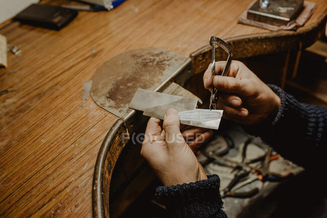 Manos de joyero anónimo usando una pinza para medir el grosor de una lámina de metal mientras se hacen joyas en el taller - foto de stock