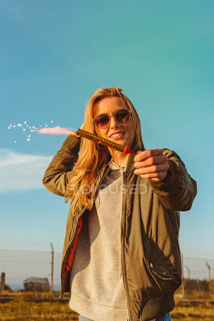Low angle of smiling blond female in sunglasses holding glowing sparkling candle in countryside with blue sky — Stock Photo