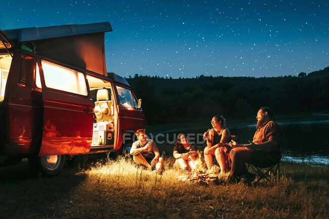 Alegre grupo de amigos que se divierten acampando cerca del lago y la camioneta durante la noche estrellada - foto de stock