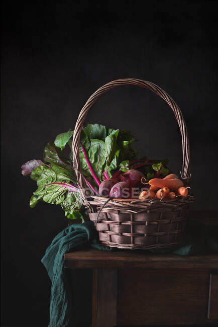 Still life of a basket with vegetables on a wooden table on a black background — Stock Photo