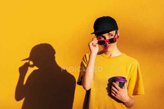 Young informal female in trendy cap sipping takeaway hot drink against yellow background — Stock Photo