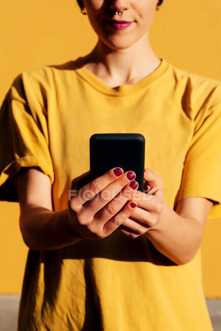 Сучасна жінка зі стильною стрижкою та пірсингом використовує смартфон у соціальних мережах на жовтому тлі — стокове фото