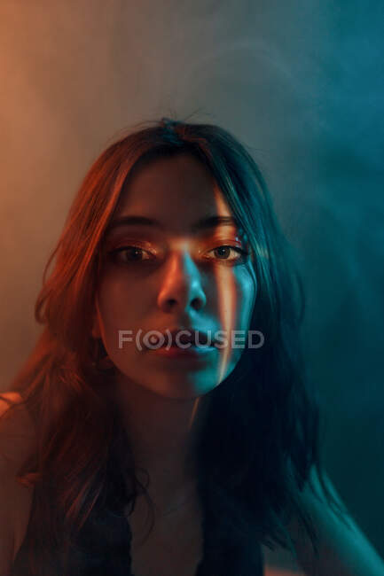 Joven modelo femenino sin emociones con proyección de luz en la cara sentado en un estudio oscuro y mirando a la cámara - foto de stock