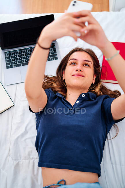Зверху позитивна молода студентка лежить на ліжку і приймає селфі на смартфон, маючи перерву під час дистанційного онлайн-навчання вдома — стокове фото
