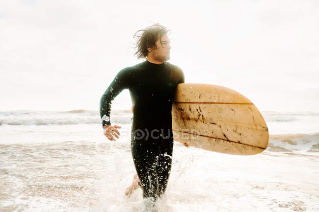 Surfer uomo vestito in muta correre via guardando con la tavola da surf sulla spiaggia durante l'alba — Foto stock