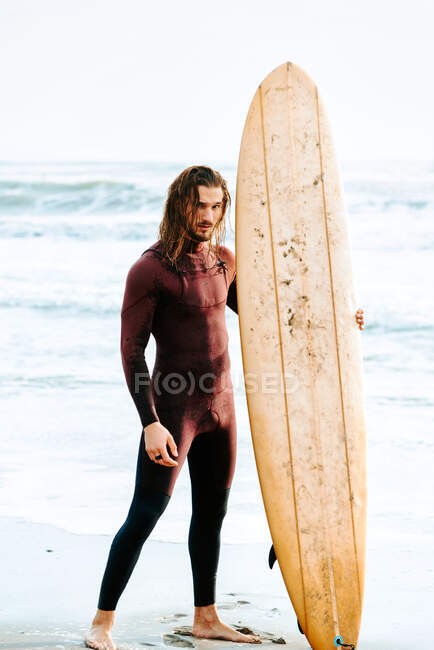 Joven surfista hombre de pelo largo vestido con traje de neopreno de pie mirando a la cámara con tabla de surf hacia el agua para coger una ola en la playa durante el amanecer - foto de stock