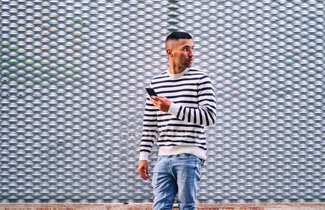 Доросла людина в повсякденному одязі стоїть біля сірої декоративної стіни і переглядає соціальні медіа на смартфоні на вулицях міста. — стокове фото