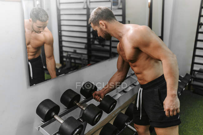 Мужчина берет тяжелые гантели со стойки во время занятий тяжелой атлетикой в спортзале — стоковое фото