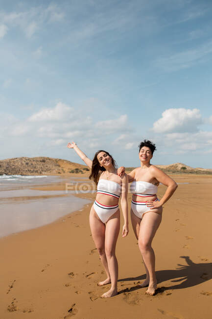 Full body of cheerful girlfriends in swimwear standing on sandy beach washed by waving sea — Stock Photo
