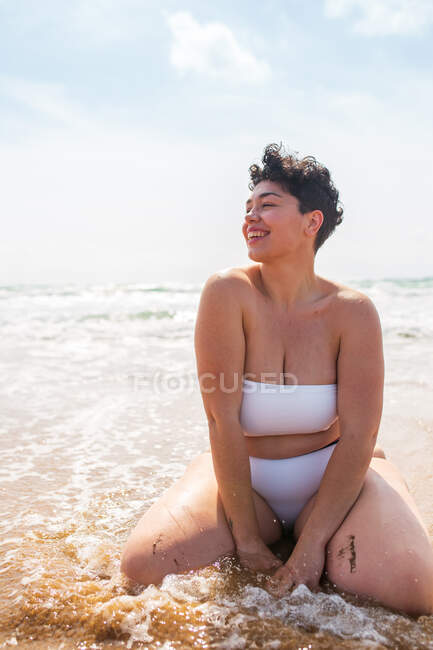 Sorrindo jovem plus size fêmea em maiô sentado na praia de areia olhando para longe perto do oceano espumoso sob céu azul nublado à luz do dia — Fotografia de Stock