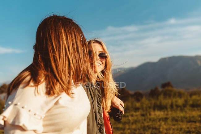 Молоді усміхнені подружки з фотоапаратом дивляться один на одного і обіймаються проти блакитного неба в полі — стокове фото