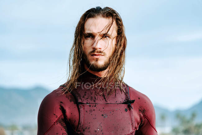 Giovane bel surfista uomo con i capelli lunghi vestito in muta in piedi guardando lontano sulla spiaggia durante l'alba — Foto stock