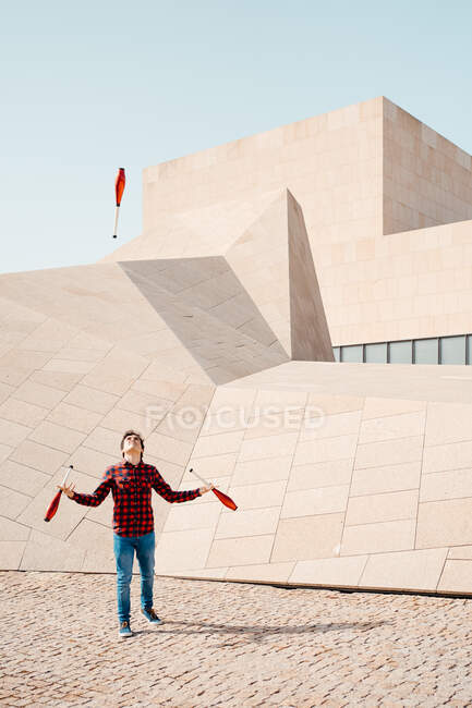 Повна довжина чоловіків, які виконують трюк з жонглювання клубами, стоячи проти сучасної кам'яної будівлі з незвичайною геометричною архітектурою — стокове фото
