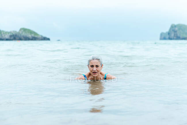 Nuoto femminile anziano dai capelli grigi attivo nelle acque oceaniche mentre si gode l'estate e pratica uno stile di vita sano sulla riva del mare — Foto stock