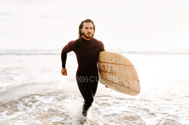 Hombre surfista vestido con traje de neopreno corriendo con tabla de surf en la playa durante el amanecer - foto de stock