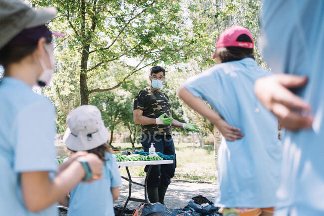 Male activist in protective mask and gloves explaining task to group of kids collecting garbage during environmental campaign in summer forest — Stock Photo