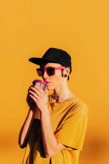 Joven mujer informal en gorra de moda sorbiendo bebida caliente para llevar contra fondo amarillo - foto de stock